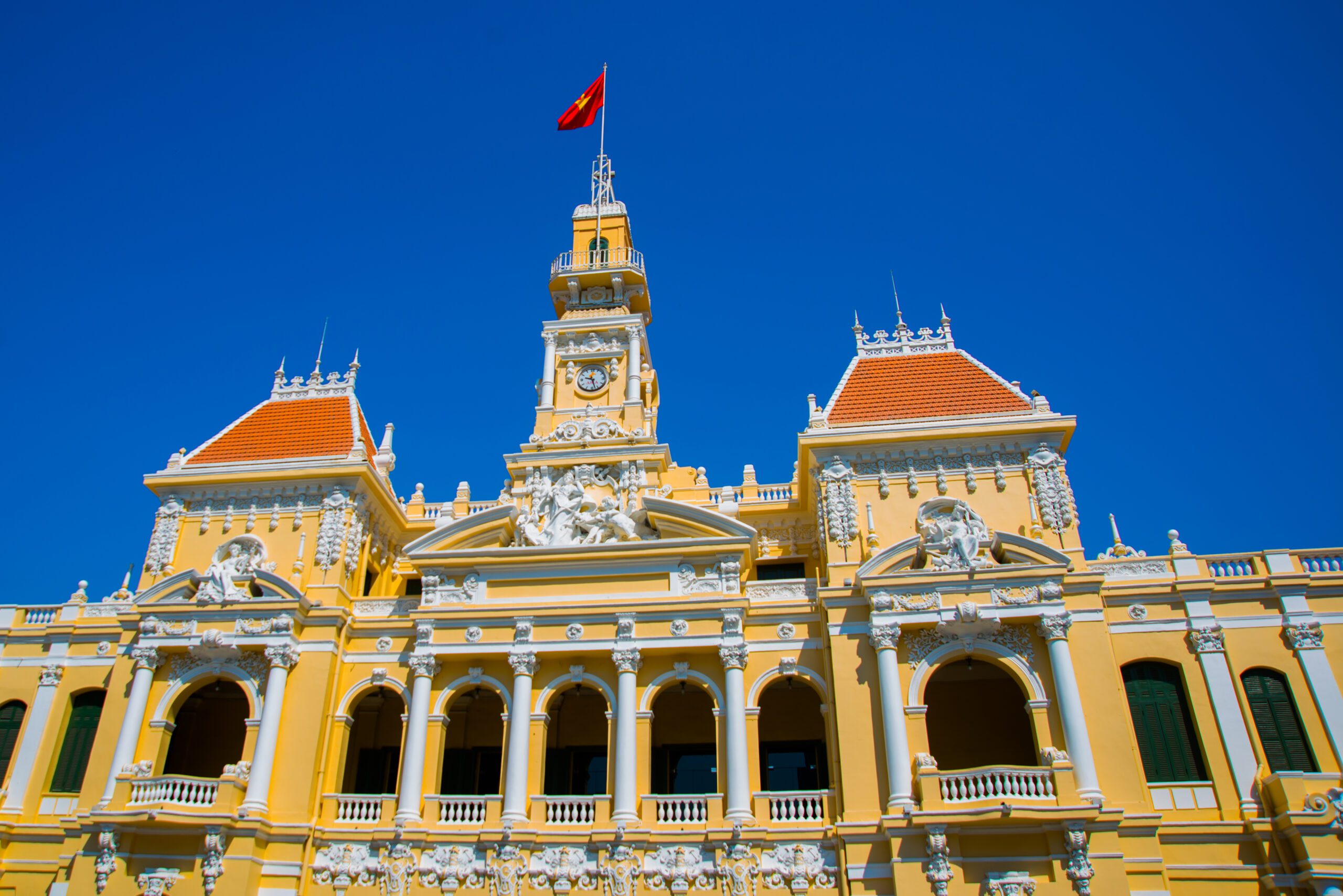French style of building in Vietnam, Asia. Beautiful Ho Chi Minh City Hall. Facade of house with ornate design. Red flag contrasts with blue sky and clouds. Tourist attraction, famous landmark.Asia